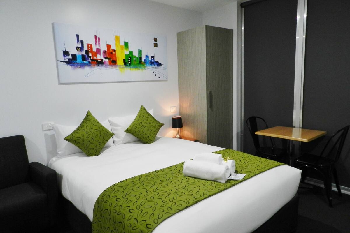 A hotel room with a bed, green patterned bedspread, two pillows, a nightstand with a lamp, a small table, and a chair.