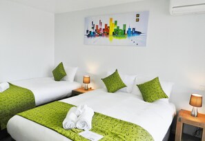 Une chambre d’hôtel avec deux lits, une literie à motifs verts, des oreillers blancs et un tableau coloré représentant un paysage urbain accroché au mur.