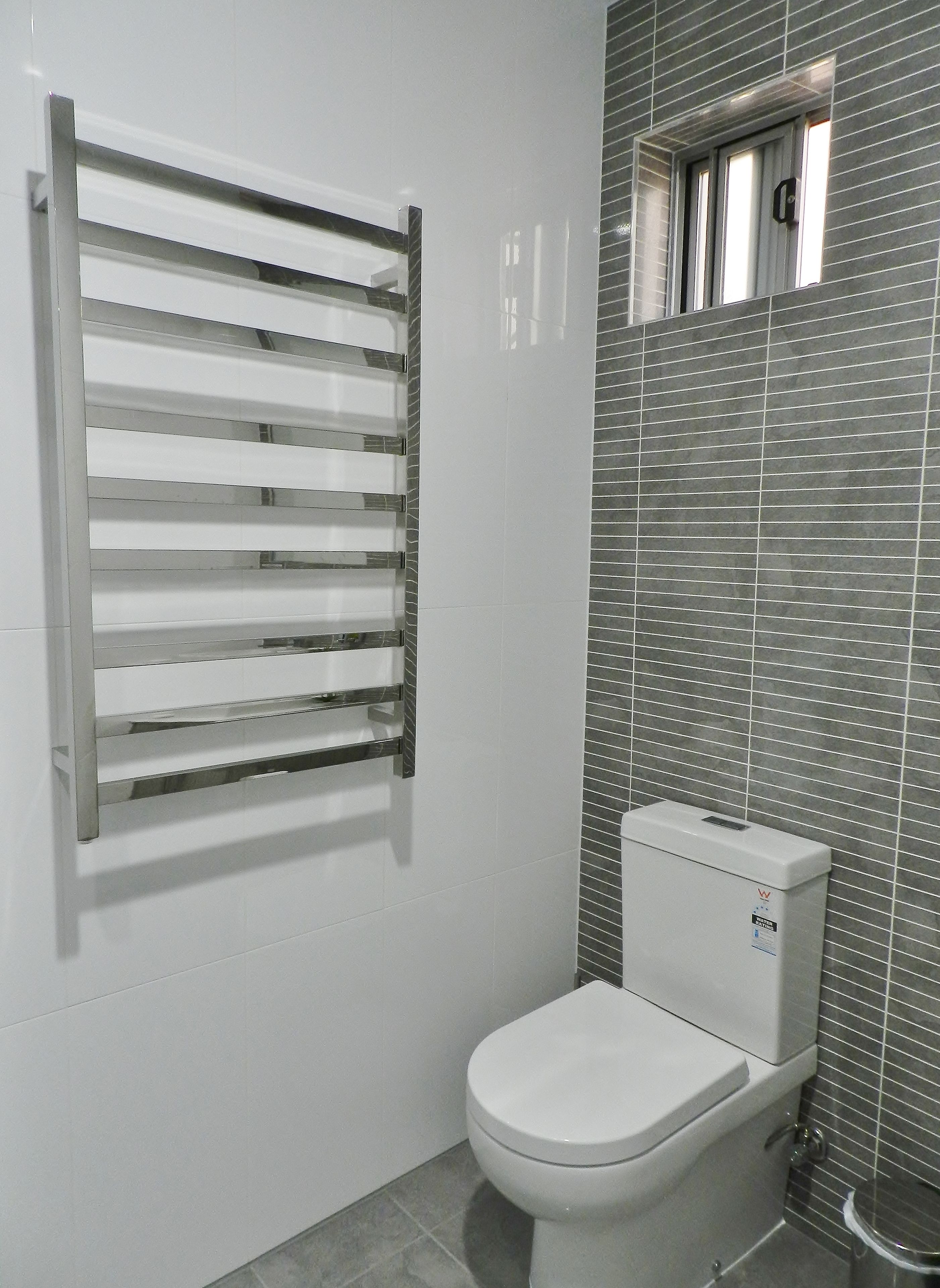 A bathroom with a white toilet, a chrome towel rack, and a tiled wall with a window.