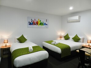 Ett hotellrum med två sängar, grönt sänglinne, vita kuddar, en tavla med ett stadsmotiv, en luftkonditioneringsanläggning och ett skrivbord med en stol.