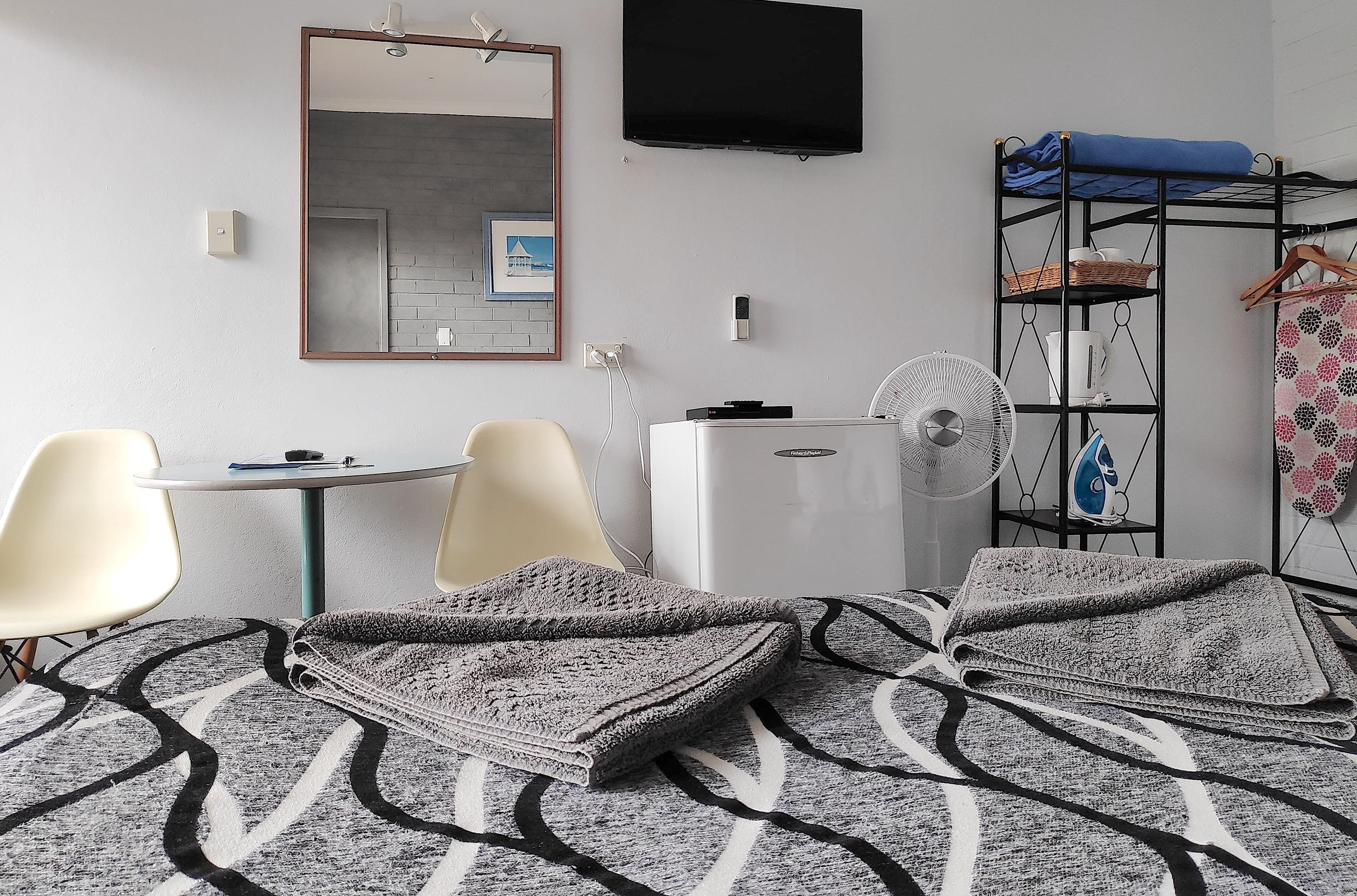 A hotel room with a bed, two chairs, a small table, a TV, a heater, a fan, and a wardrobe with a hanging rack.