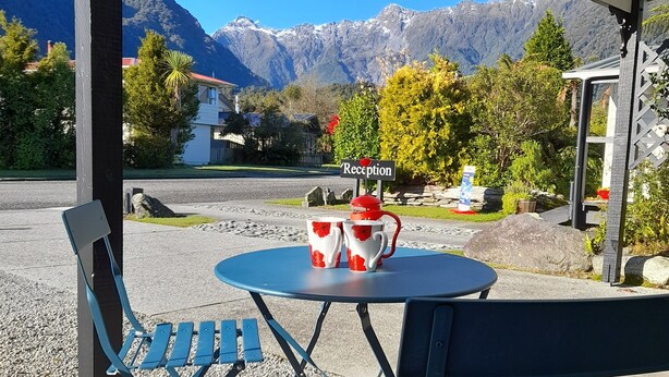 Un'area salotto esterna riparata con tavolo e sedie, una targa con la reception e vista sulle montagne sullo sfondo.