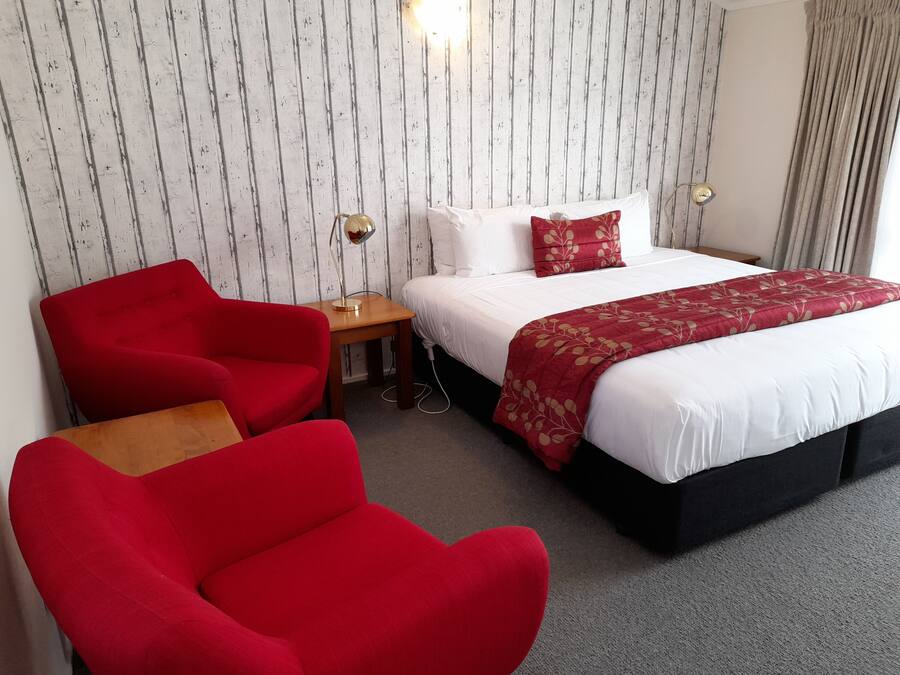 A hotel room with a bed, two red chairs, a small table, and a lamp.