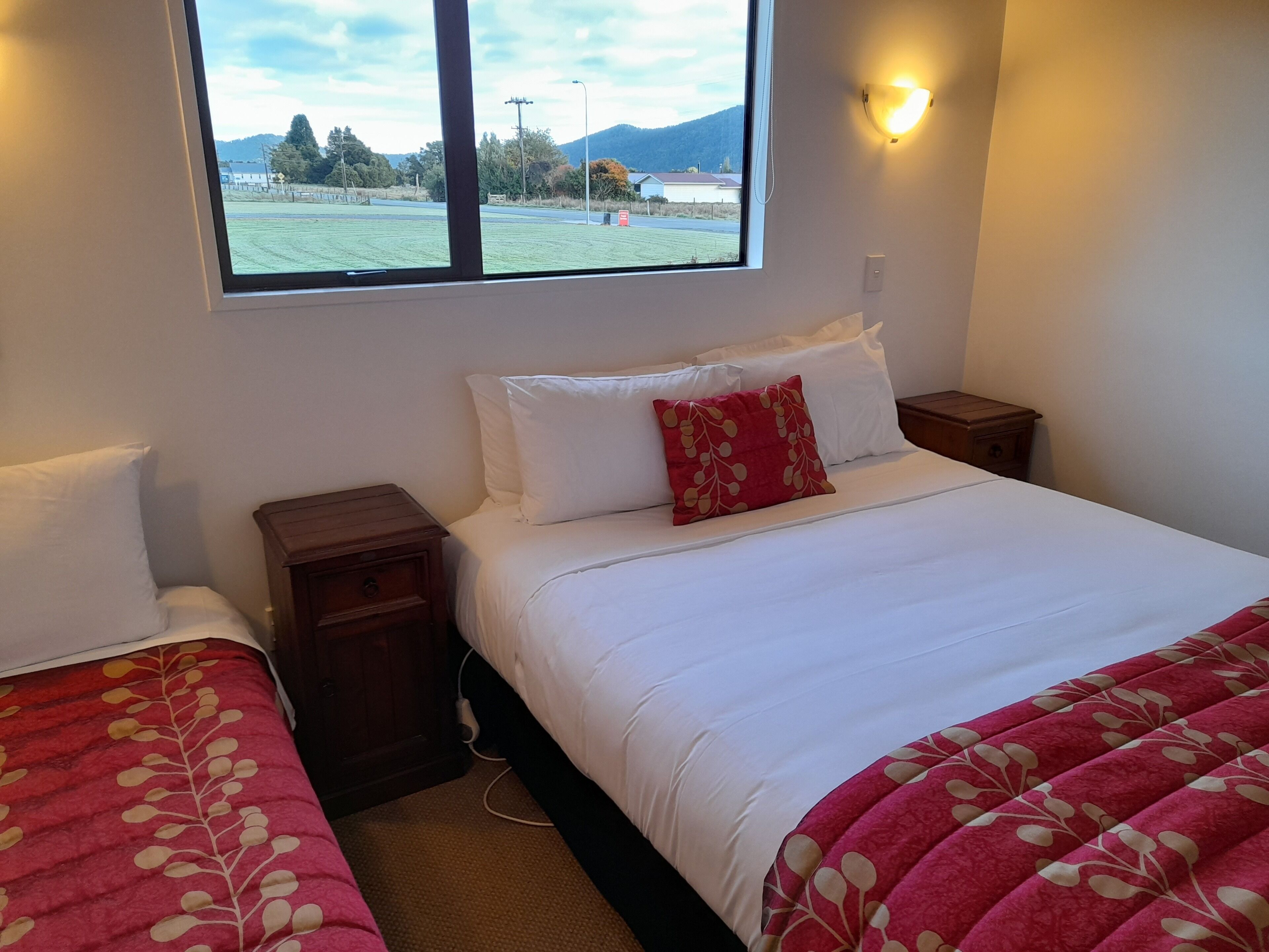 Un dormitorio con una cama, dos mesitas de noche, un ventanal con vistas a un campo y montañas, y una lámpara fijada a la pared.