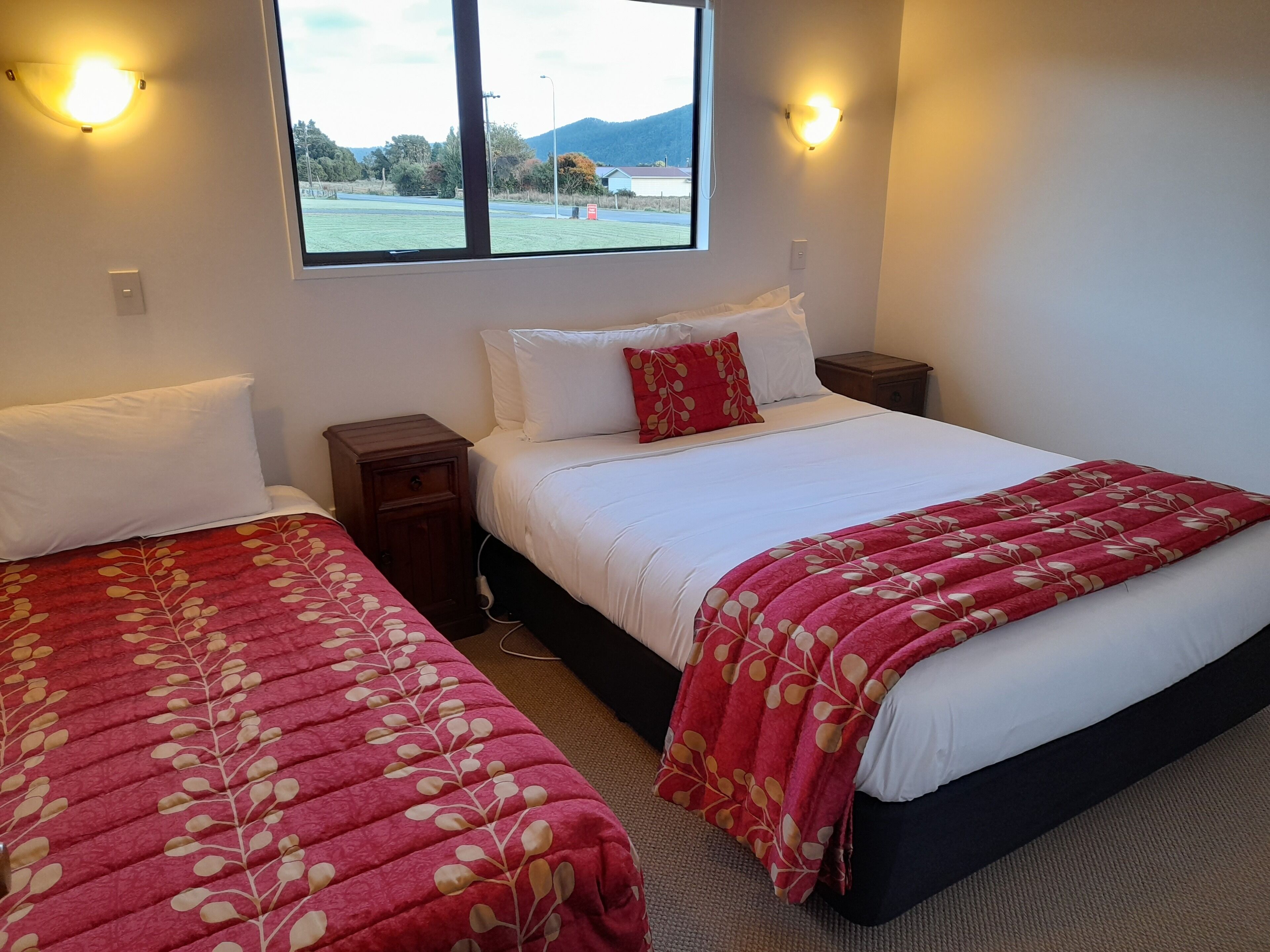 A bedroom with two beds, each with a red floral bedspread, white pillows, and a window with a view of a landscape.