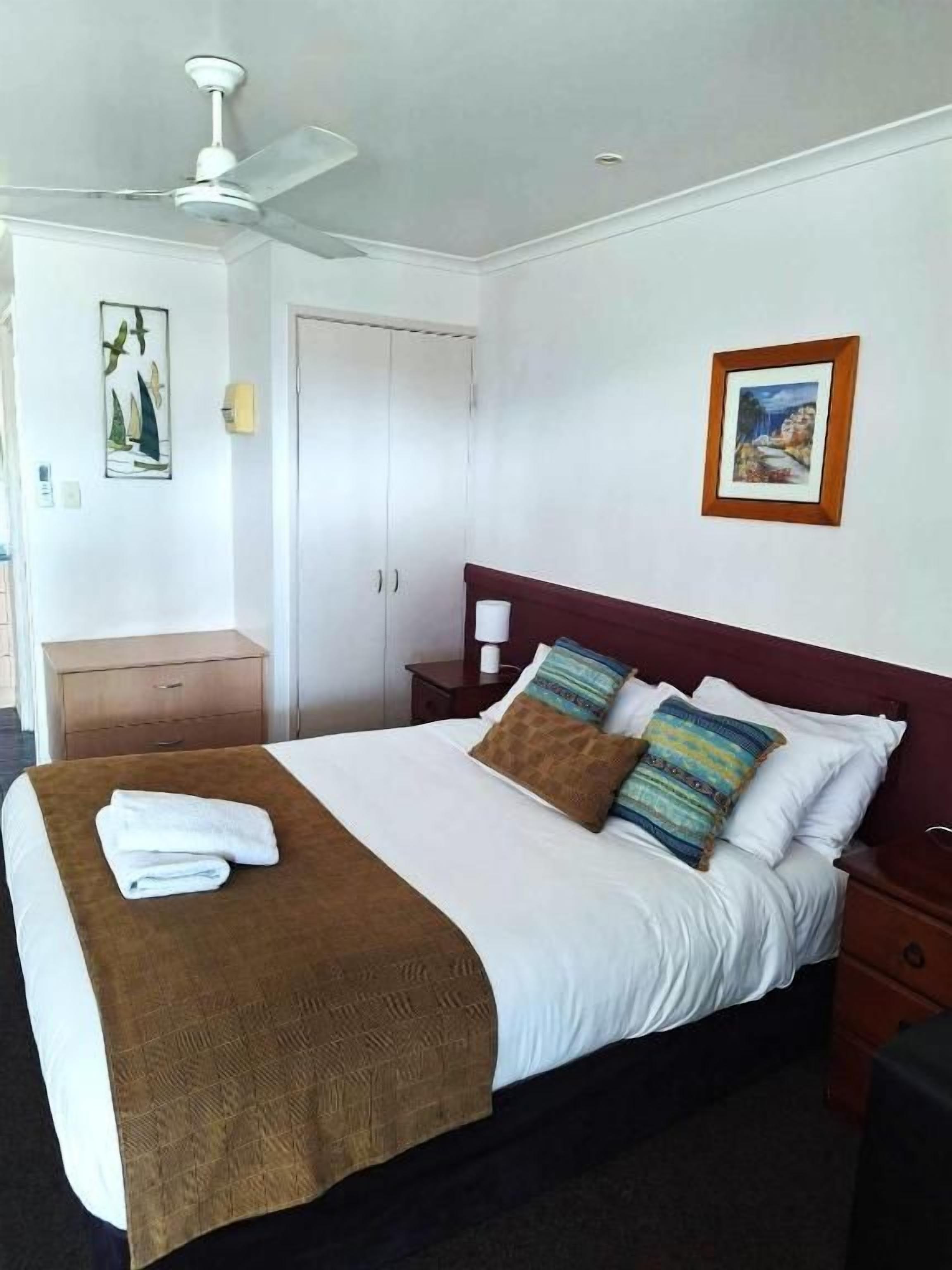 A hotel room with a bed, a nightstand, a wardrobe, a ceiling fan, and two framed pictures on the wall.
