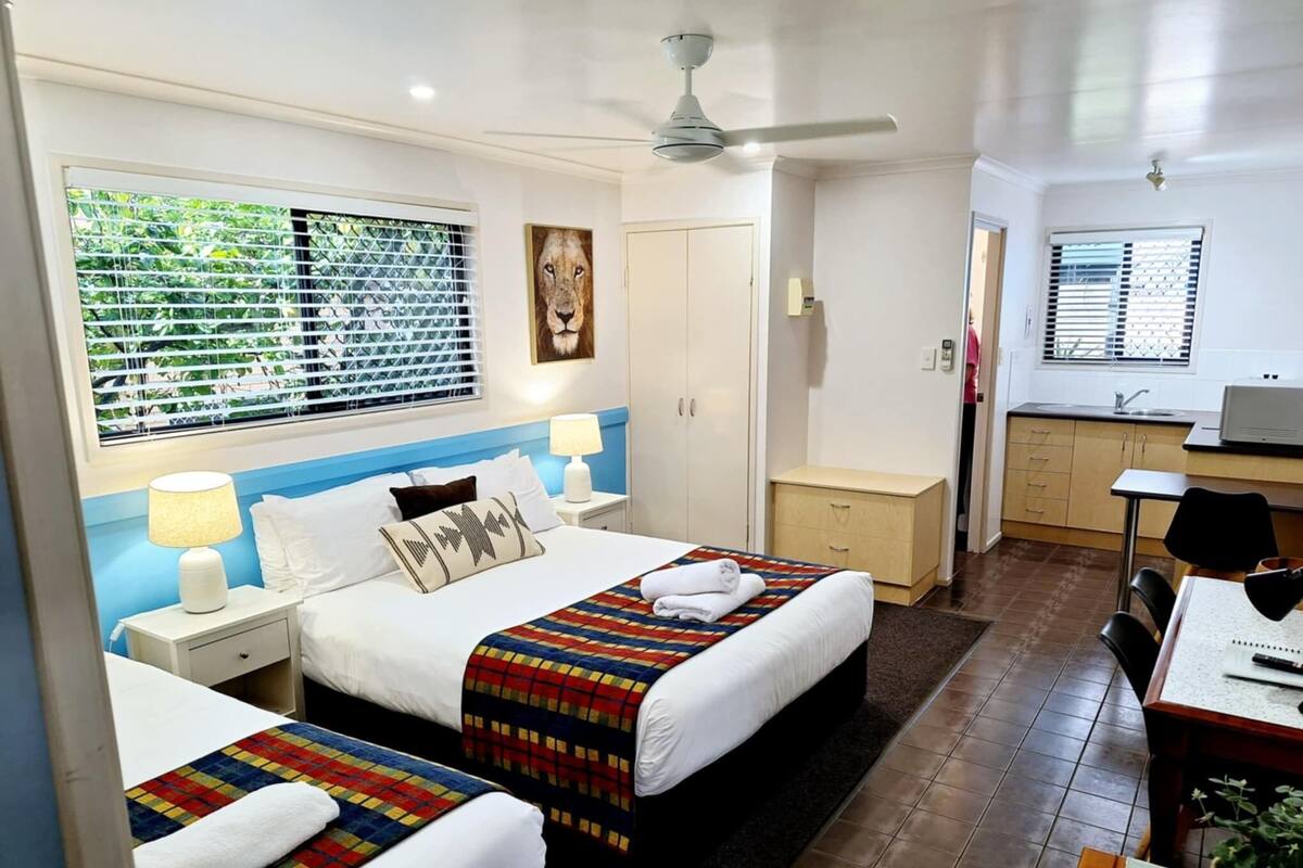 A hotel room with two beds, a desk, a chair, a ceiling fan, and a window with blinds.