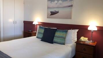 Habitación de hotel con cama, dos lámparas de noche, una mesita de noche, un teléfono y un cuadro de un barco en el agua.