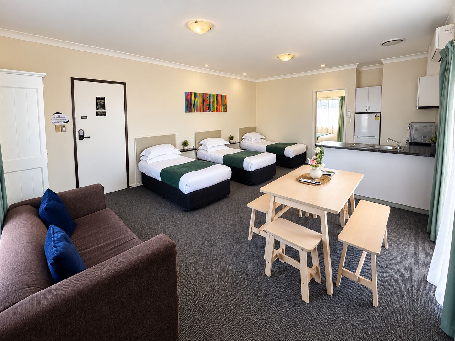 Quarto de hotel com duas camas, um sofá, uma mesa de jantar com bancos, uma cozinha compacta e um quadro na parede.