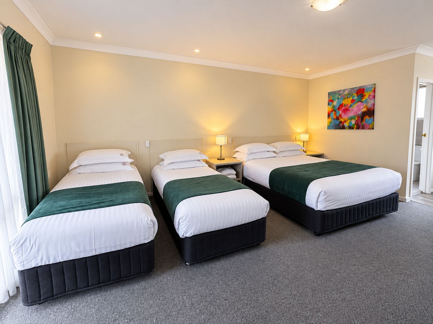 A hotel room with three beds, each with a green bedspread and white bedding. There are bedside lamps, a painting on the wall, and a door leading to another room.