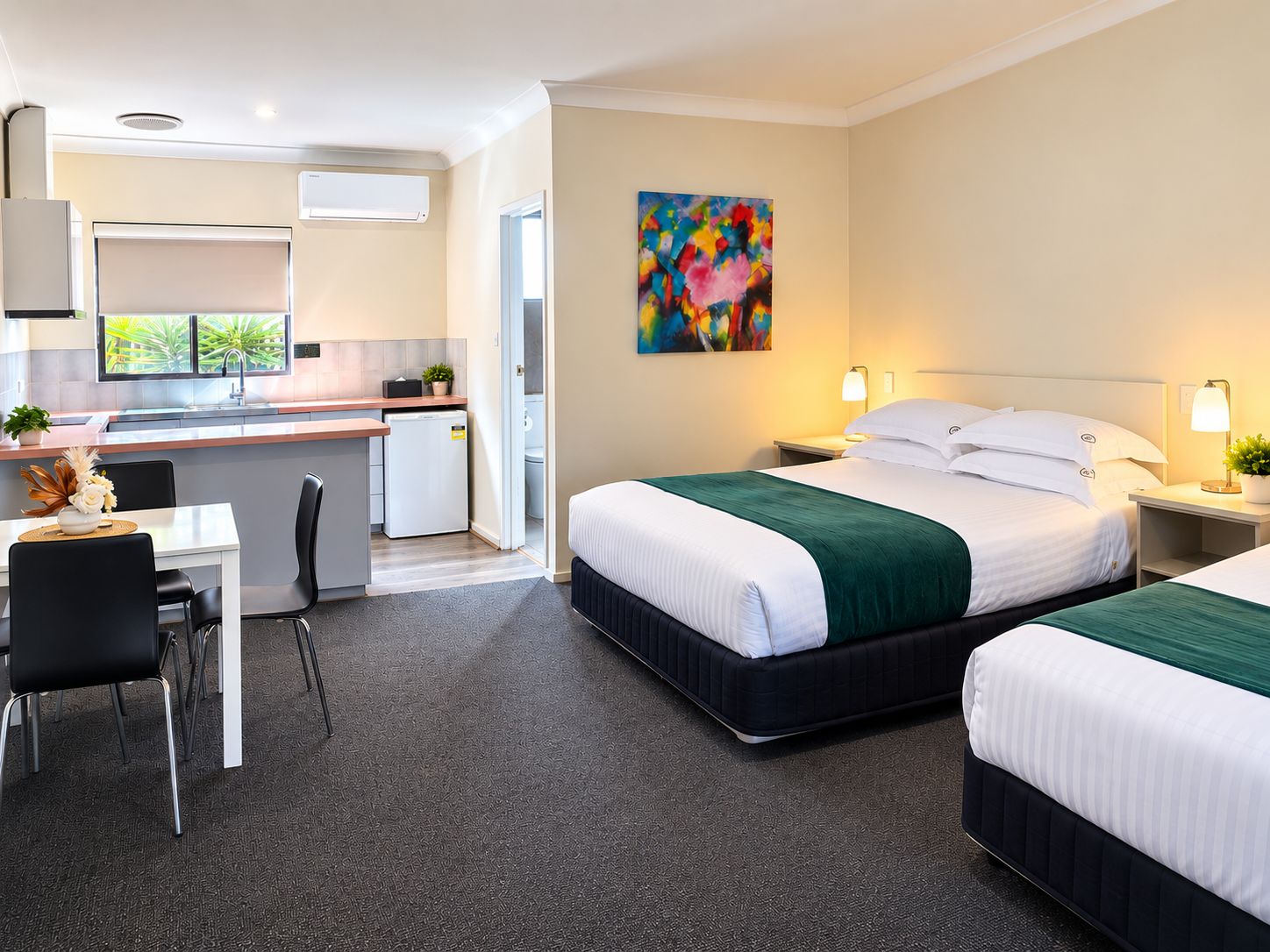 A hotel room with a bed, bedside table, lamp, and a view of a kitchen area.
