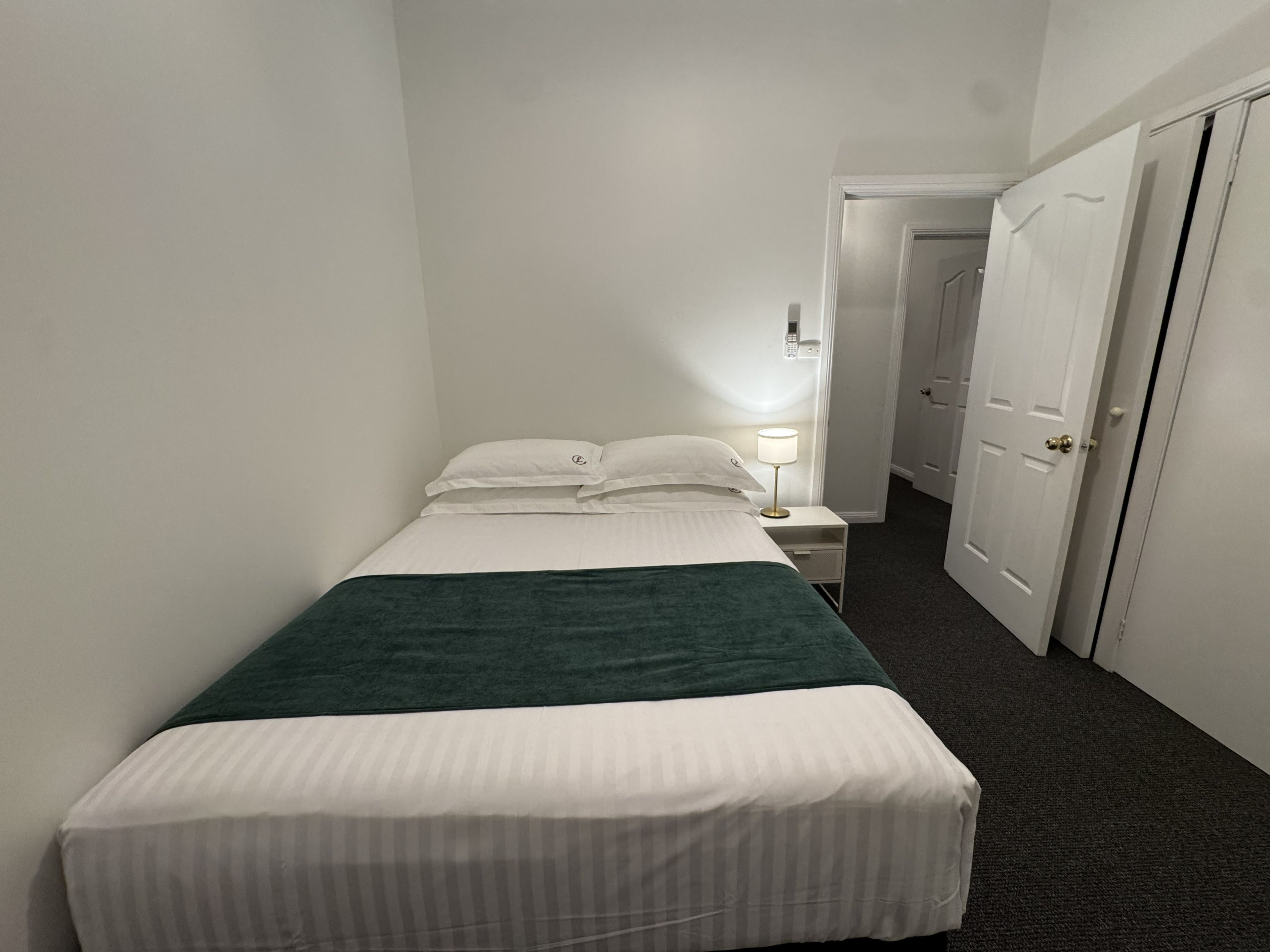 Una cama individual con una manta verde, almohadas blancas y una lámpara de noche en una habitación con paredes blancas y piso alfombrado.