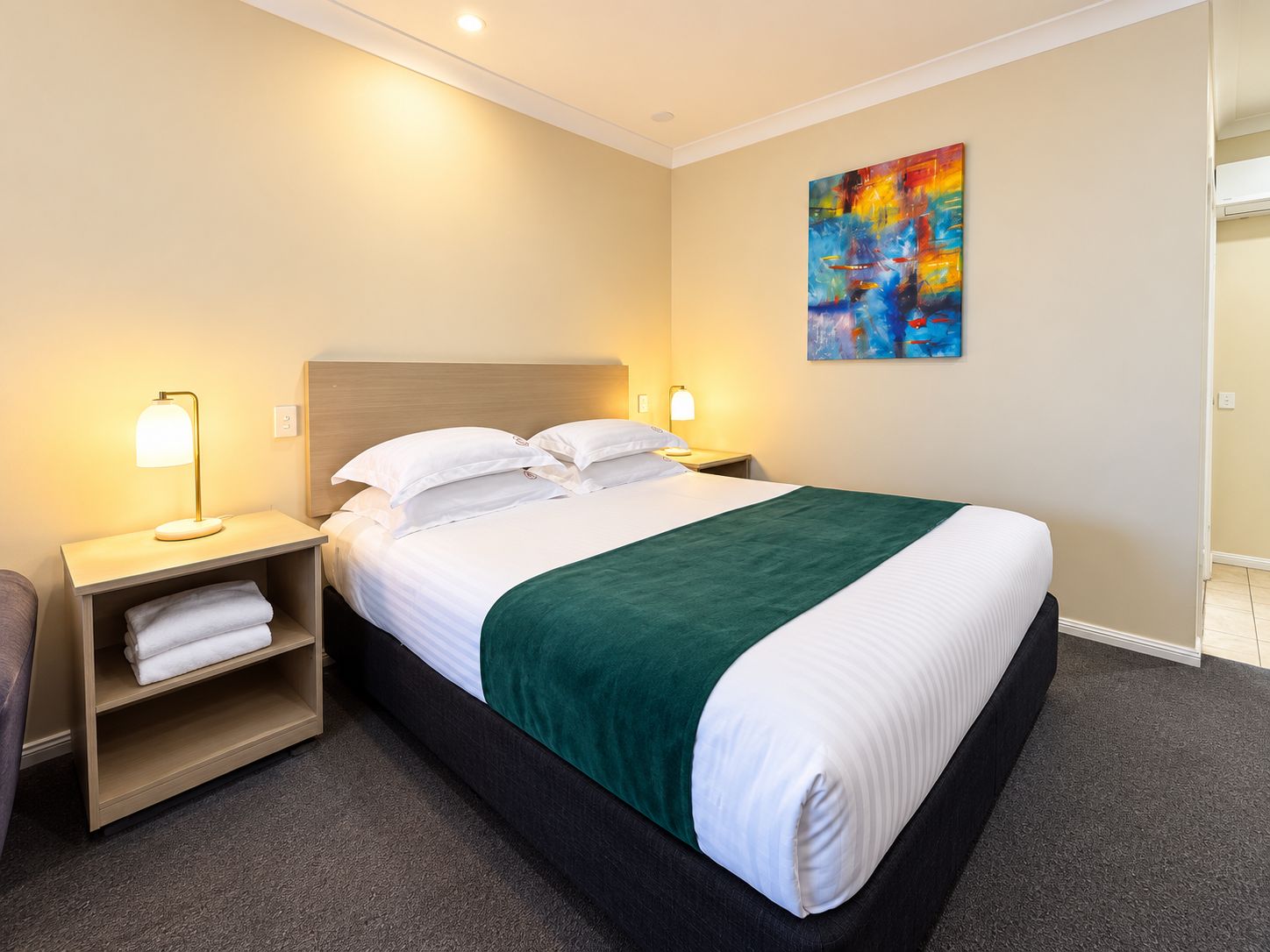 Una habitación de hotel con una cama, dos lámparas de noche, una mesita de noche con una toalla, un plaid verde y un cuadro abstracto colorido en la pared.