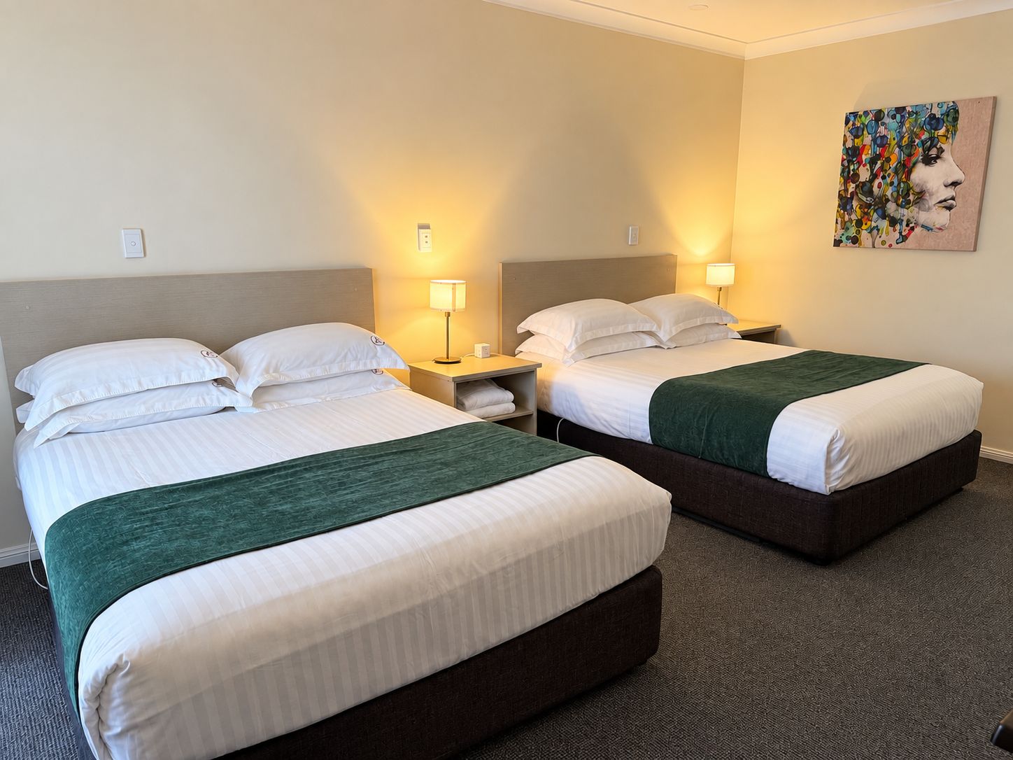 A hotel room with two beds, each with a green throw, white bedding, and bedside lamps.