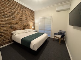 Quarto de hotel com parede de tijolos, cama com roupa de cama branca, manta verde, mesa de cabeceira, cadeira e televisão fixada na parede.