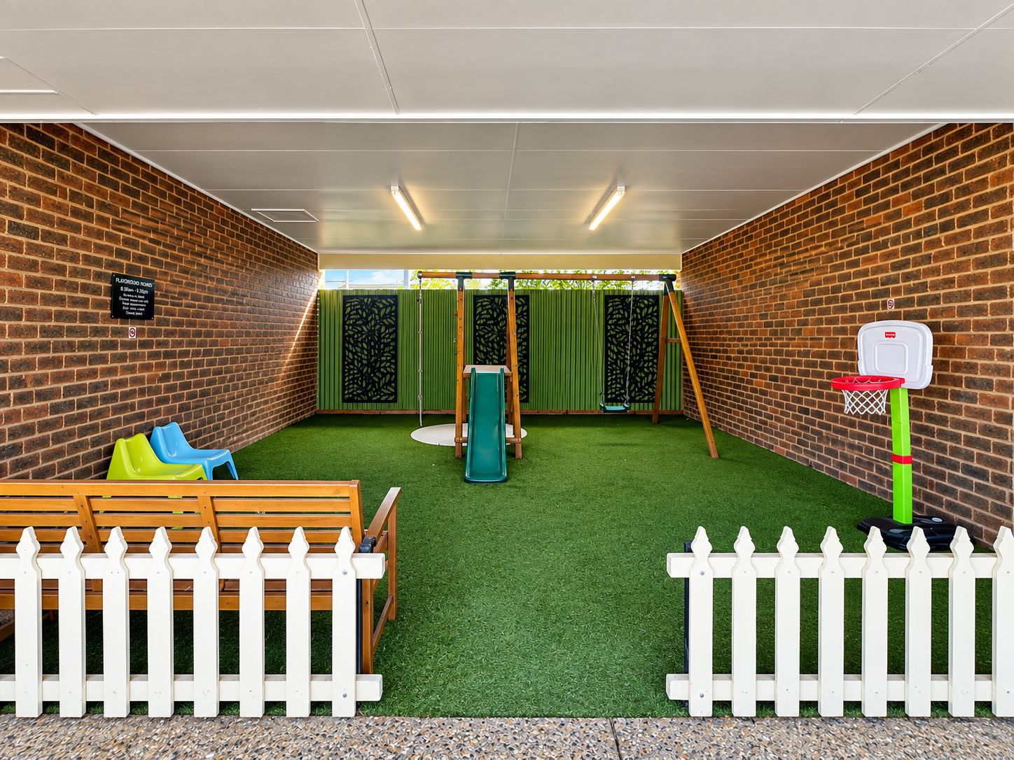 A play area with a basketball hoop, a swing set, and a slide, enclosed by a white picket fence.