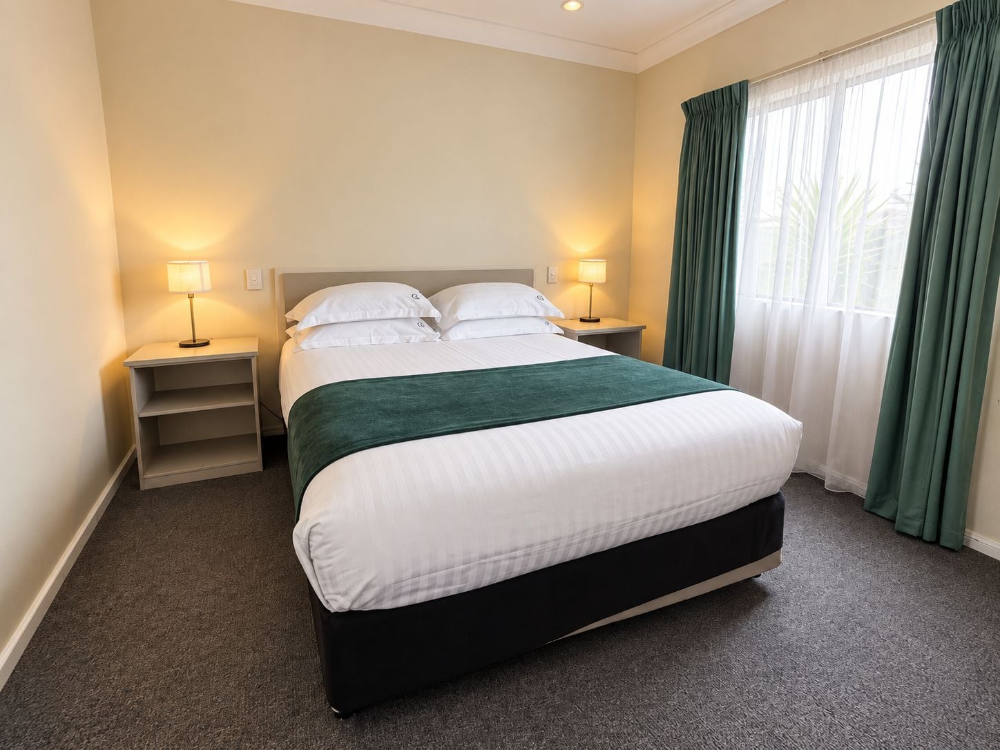 Une chambre d’hôtel avec un lit, deux tables de chevet, des lampes, un plaid vert et une fenêtre avec des rideaux.