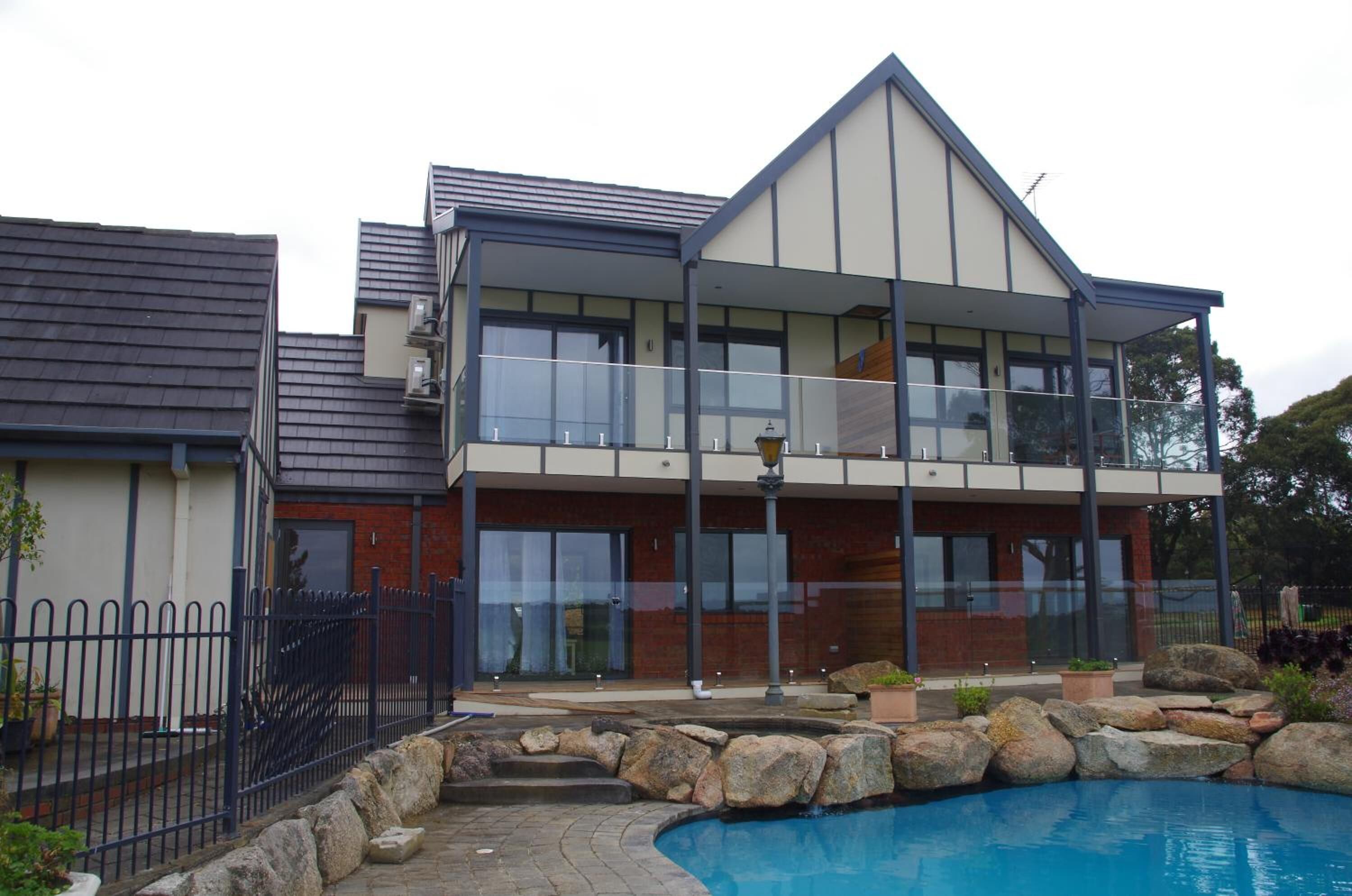 A modern two-story house with a large balcony, a swimming pool, and a stone retaining wall.