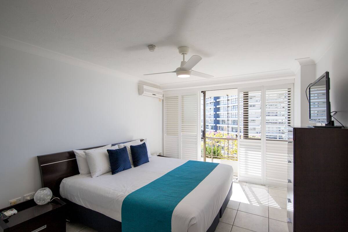 A bedroom with a bed, a nightstand, a desk, a ceiling fan, and a view of the outside.