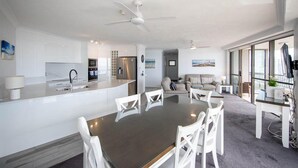 A modern kitchen with white cabinetry, a dining area with a dark table and white chairs, and a living room with a sofa and a view through large windows. - Bougainvillea Luxury Apartments (Main Beach)