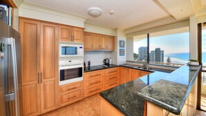 A modern kitchen with wooden cabinets, a stainless steel refrigerator, a built-in oven, and a large window offering a view of the city and ocean. - Bougainvillea Luxury Apartments (Main Beach)