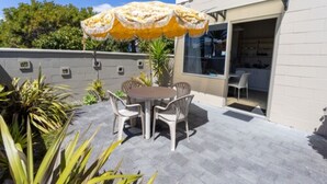 An outdoor patio with a round table and chairs, a yellow umbrella, and a glass door leading to a room. - Cosy Corner Holiday Park (Tauranga)