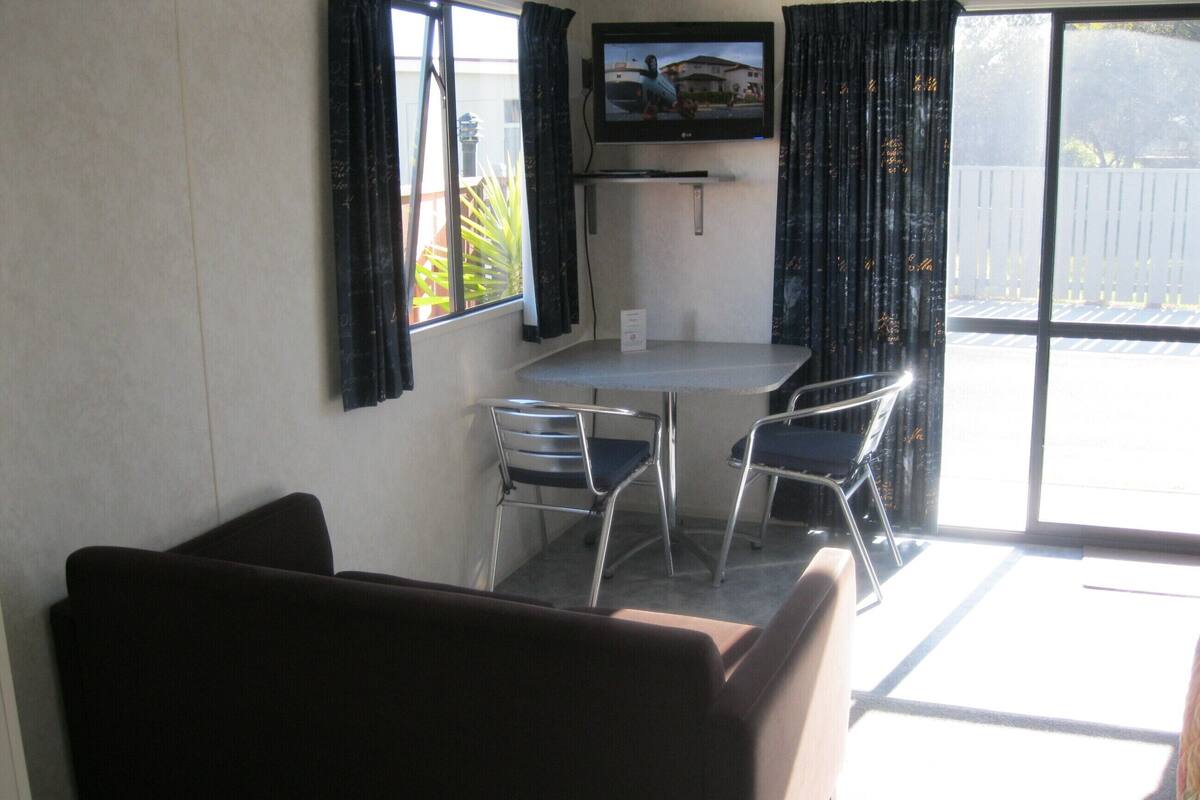 A room with a TV mounted on the wall, a table with chairs, and a large window with curtains.