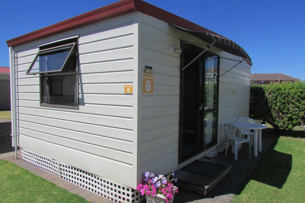A small, single-story mobile home with a red roof, a window, and a door. There is a small patio area with a table and chairs, and a flower planter with pink flowers.