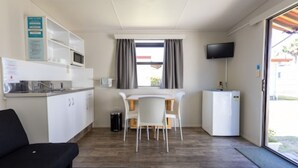 A compact hotel room with a kitchenette, a dining area with a table and chairs, a flat-screen TV mounted on the wall, and a sliding glass door leading to an outdoor area. - Cosy Corner Holiday Park (Tauranga)