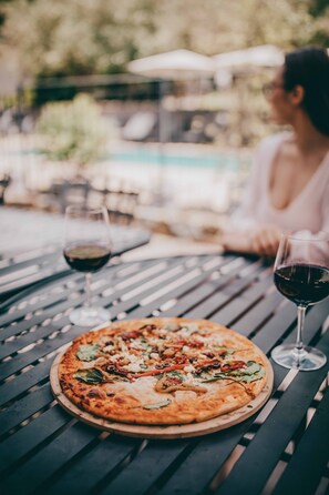 Una pizza sobre una tabla de madera, sobre una mesa de metal, con dos vasos de vino tinto.