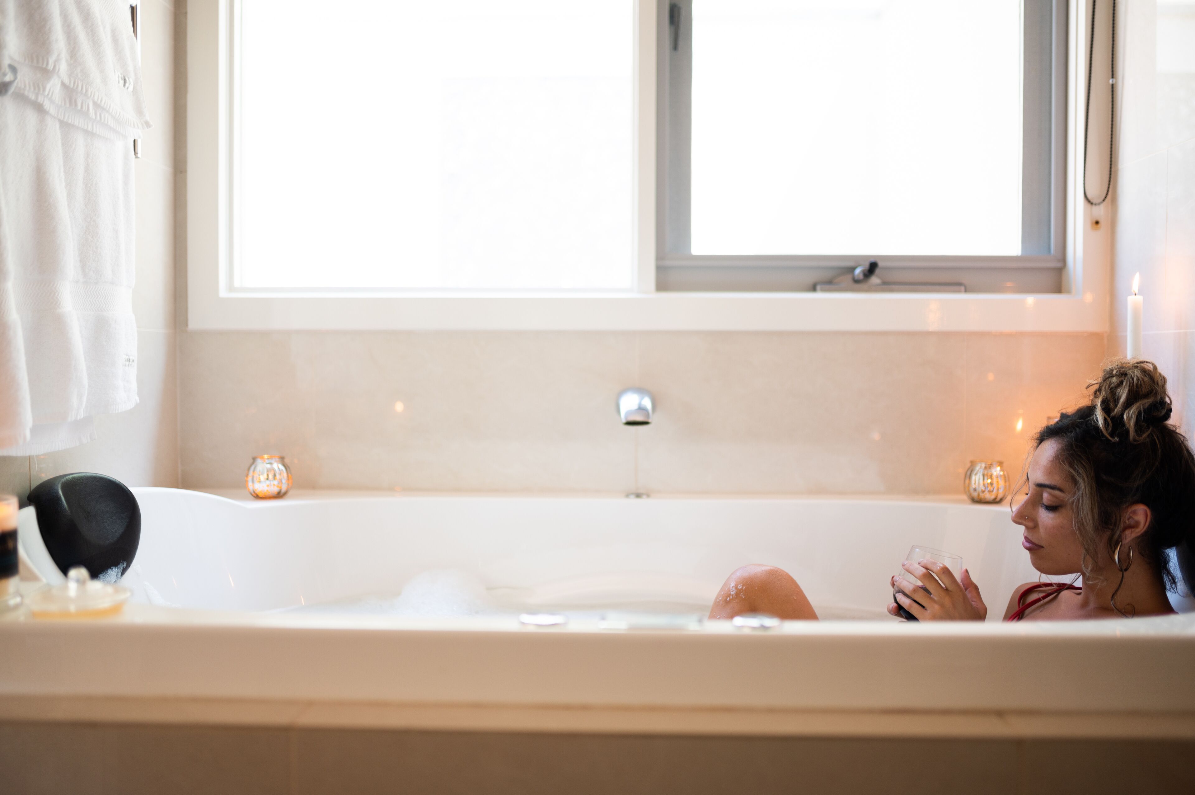 A bathtub with a window above it, a towel rack to the left, and candles on the edge of the tub.