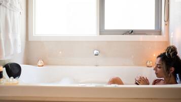 A bathtub with a window above it, a towel rack to the left, and candles on the edge of the tub.