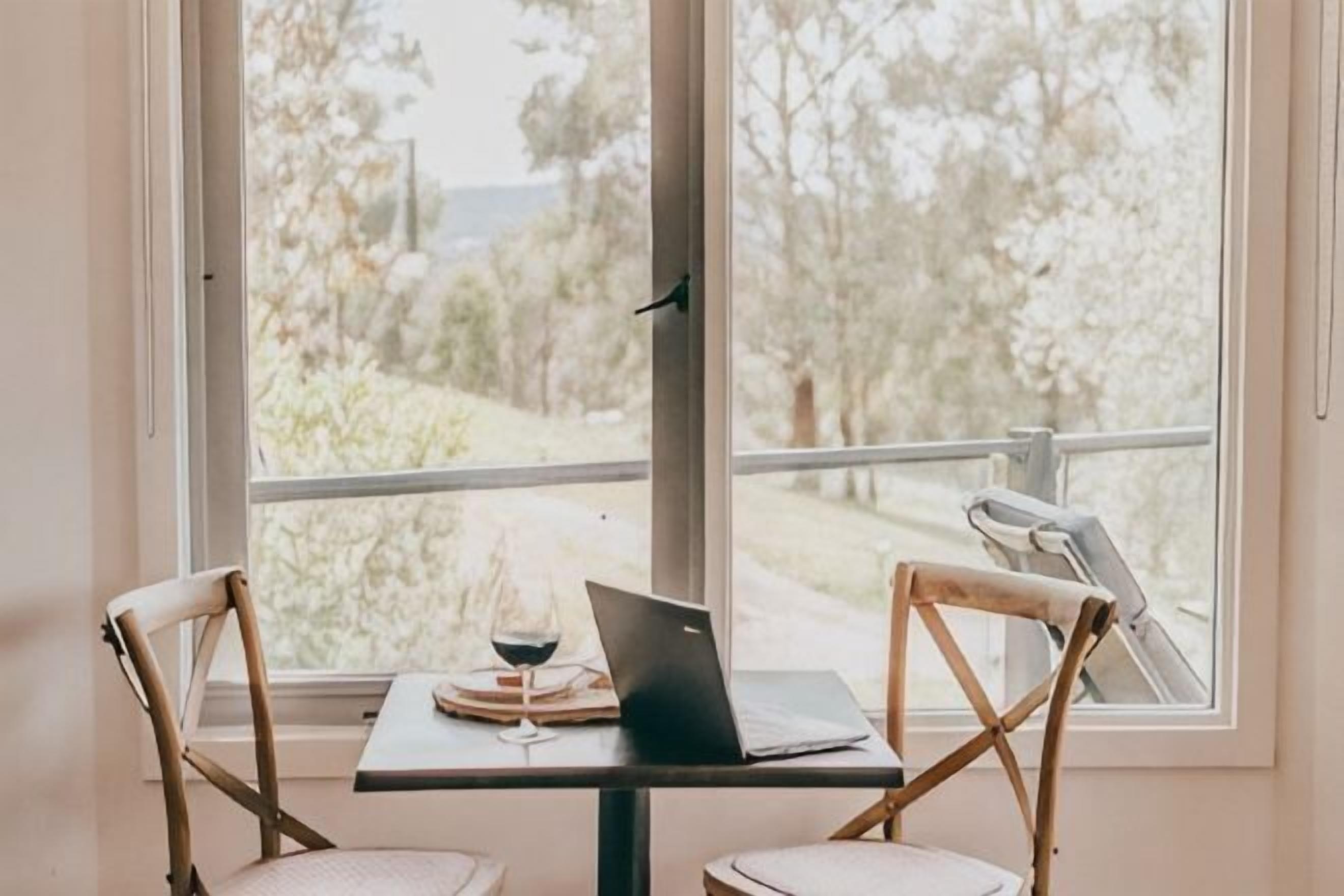 Una mesa con una computadora portátil, una copa de vino, dos sillas y vistas a través de un amplio ventanal.