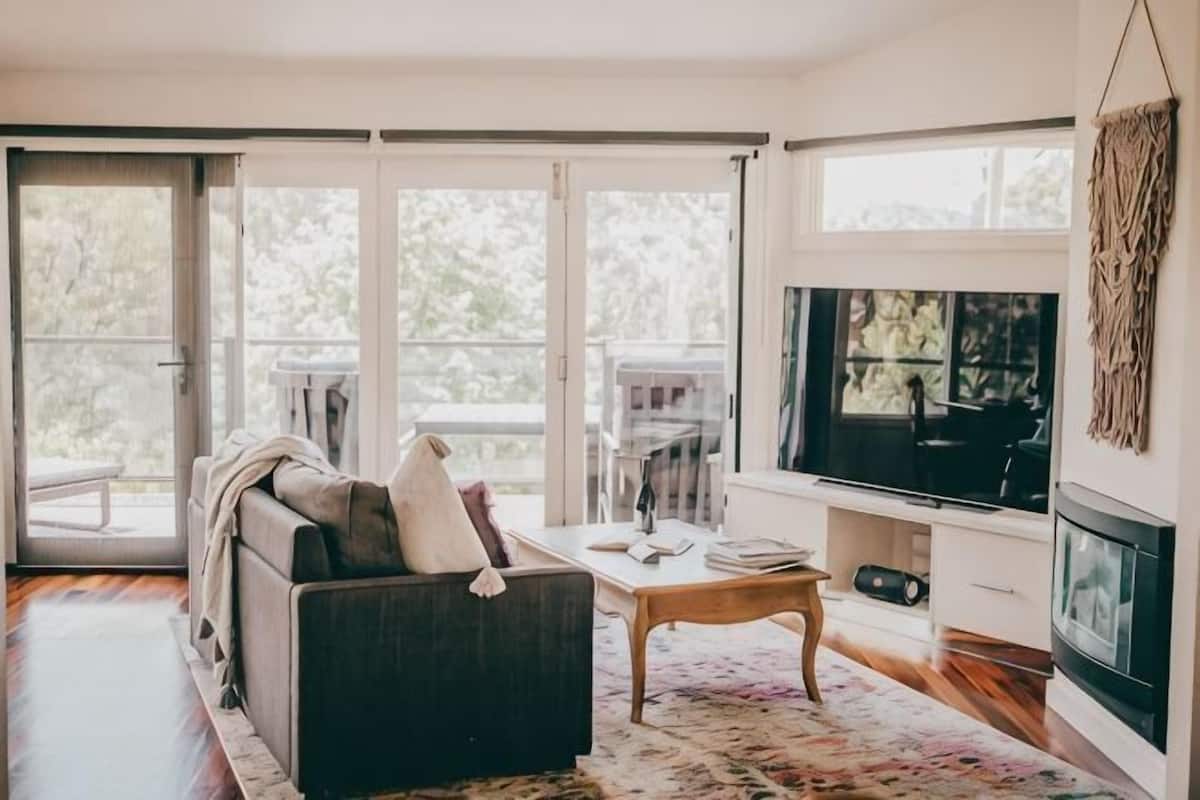 Ein Wohnzimmer mit Sofa, Couchtisch, Fernseher, Kamin und großen Fenstern.