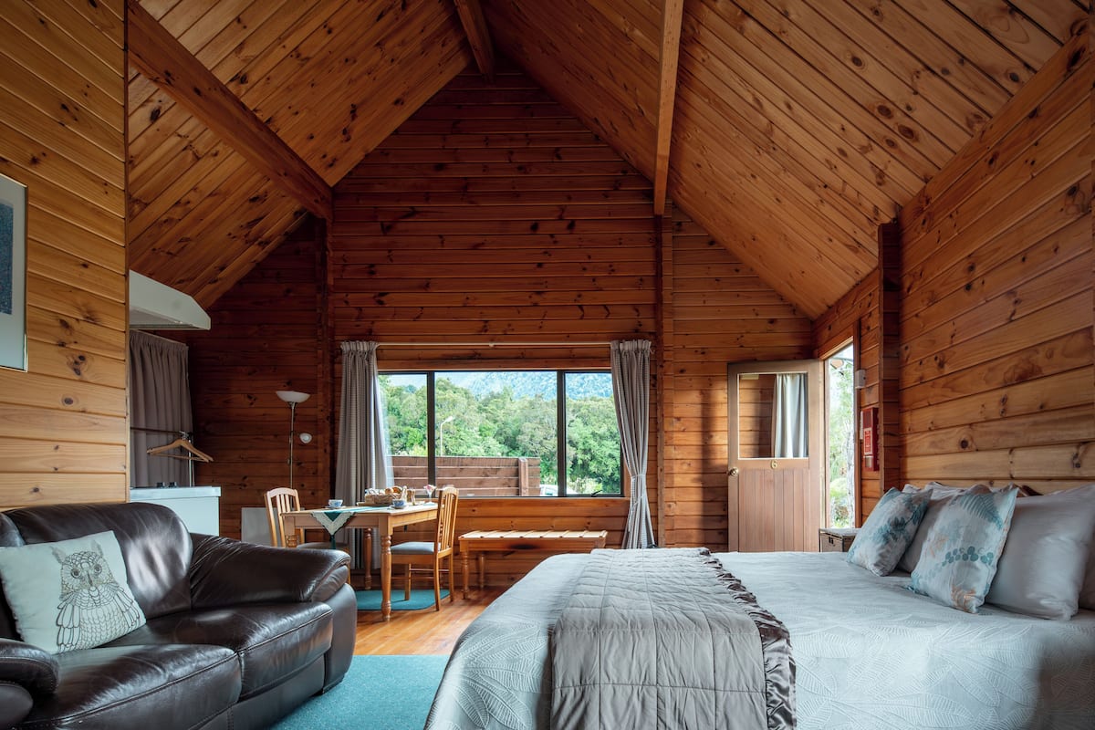 A log cabin room with a bed, a leather sofa, a dining table with chairs, and a large window offering a view of the outdoors.