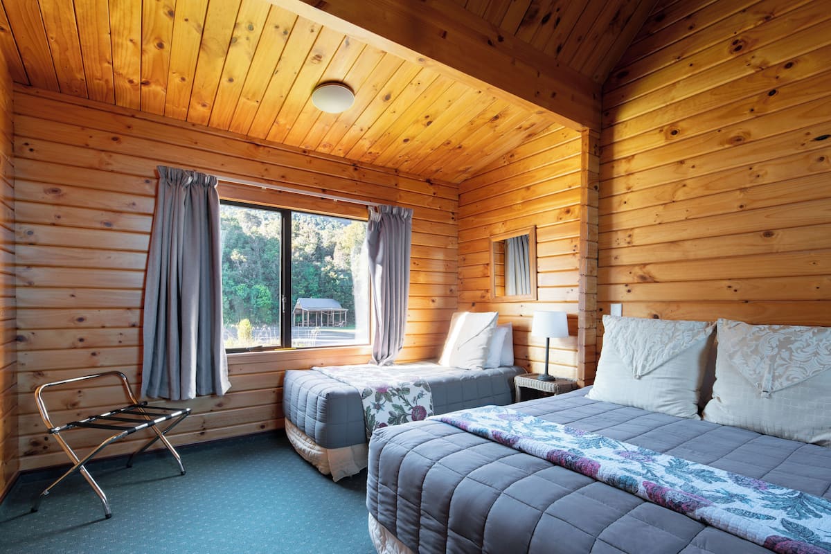 A wooden cabin room with two beds, a window with curtains, a metal chair, and a nightstand with a lamp.