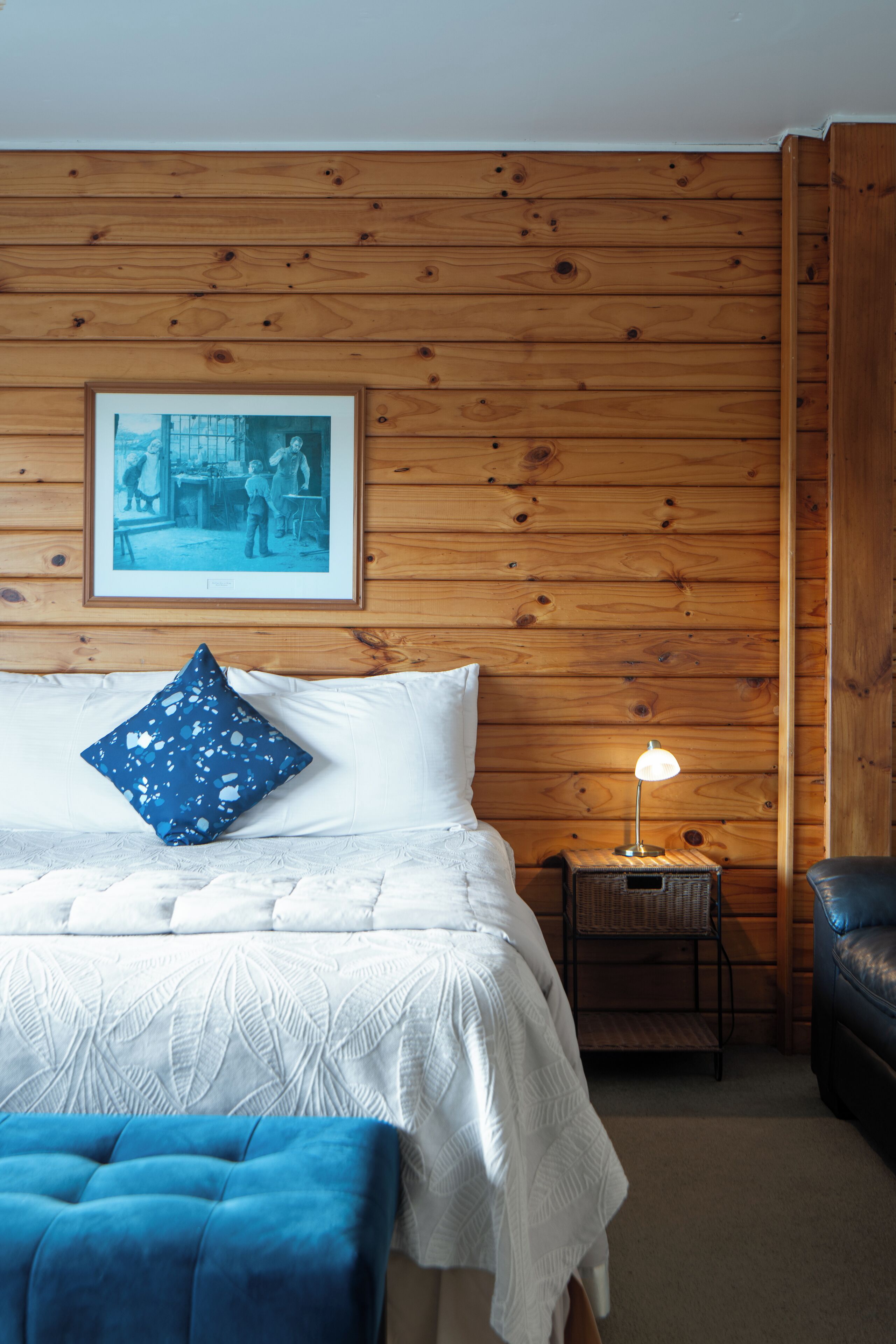 A room with a wooden wall, a bed with white bedding, a blue pillow, a small table with a lamp, and a blue chair.