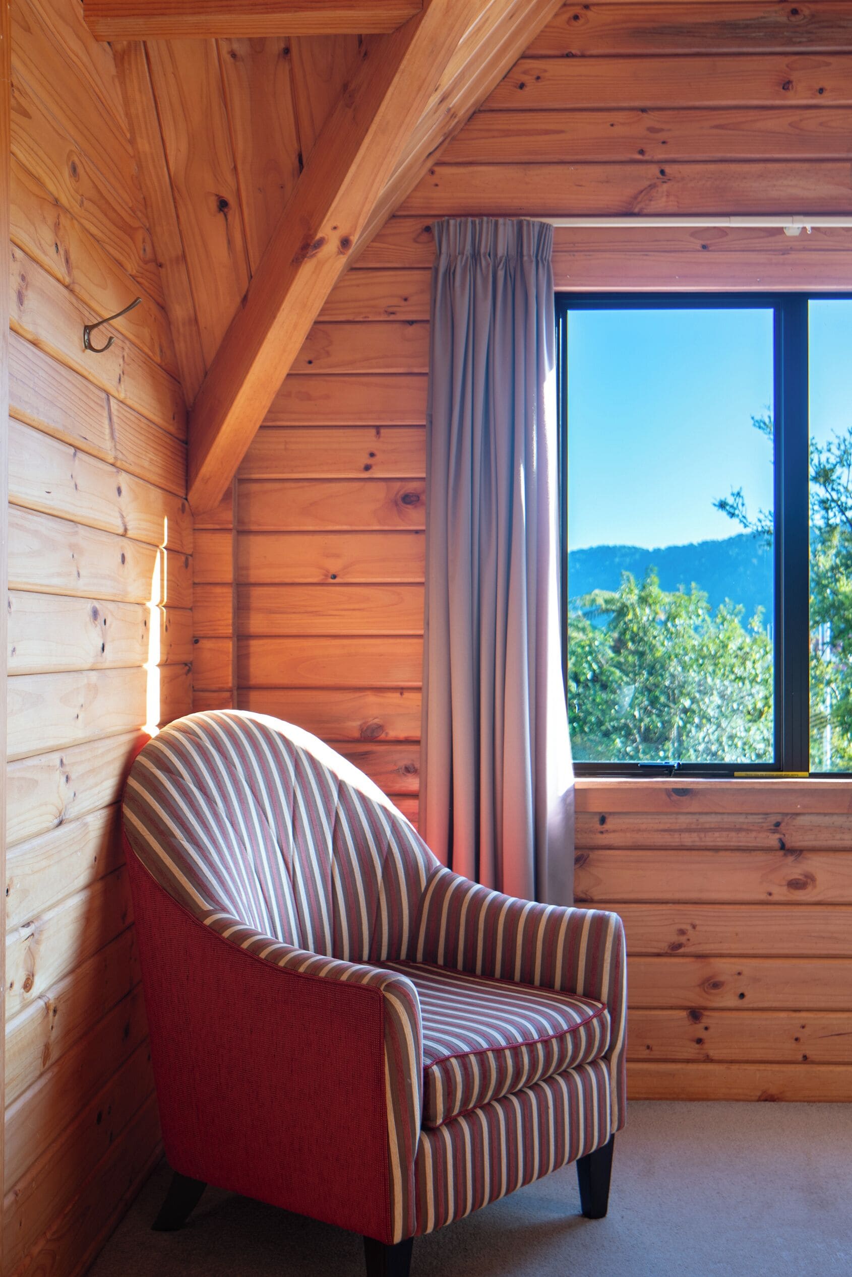 A cozy cabin room with a wooden interior, a window with a view of trees and mountains, and a striped armchair.