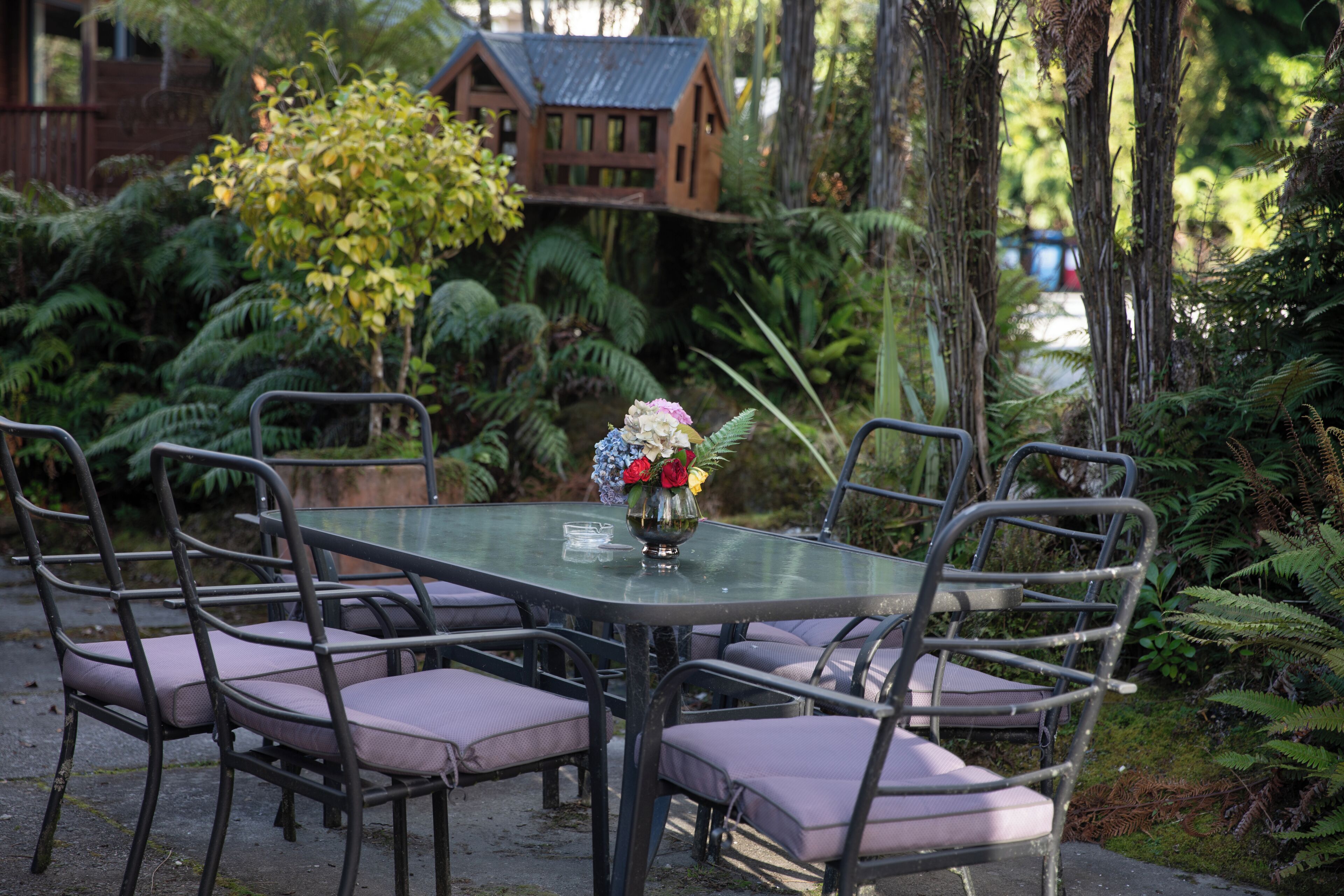 A glass-top table with six chairs, surrounded by lush greenery and a small wooden structure in the background.