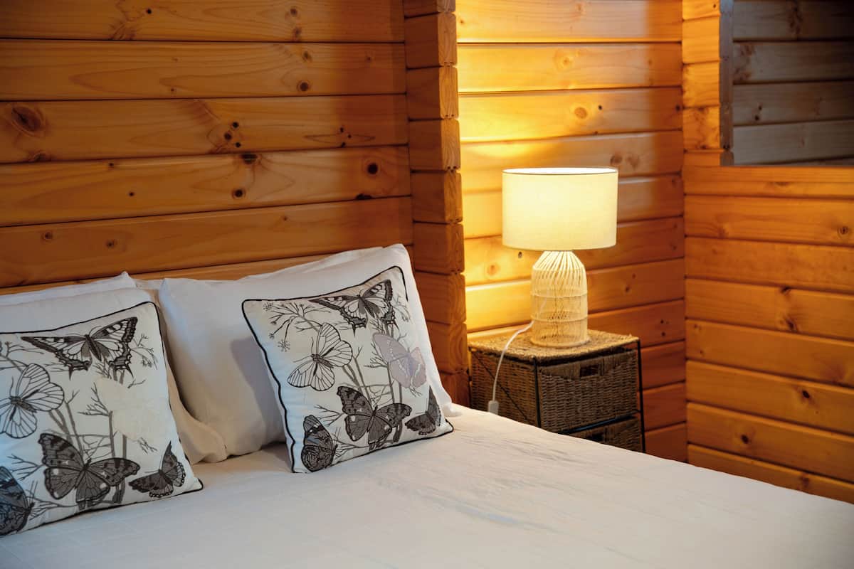 A wooden cabin room with a bed, two pillows with butterfly designs, a nightstand with a lamp, and a wicker basket.