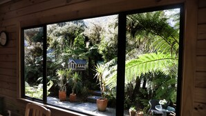 A wooden cabin with a large window overlooking a lush garden with various potted plants and a small house. - Fox Glacier Lodge (Fox Glacier)