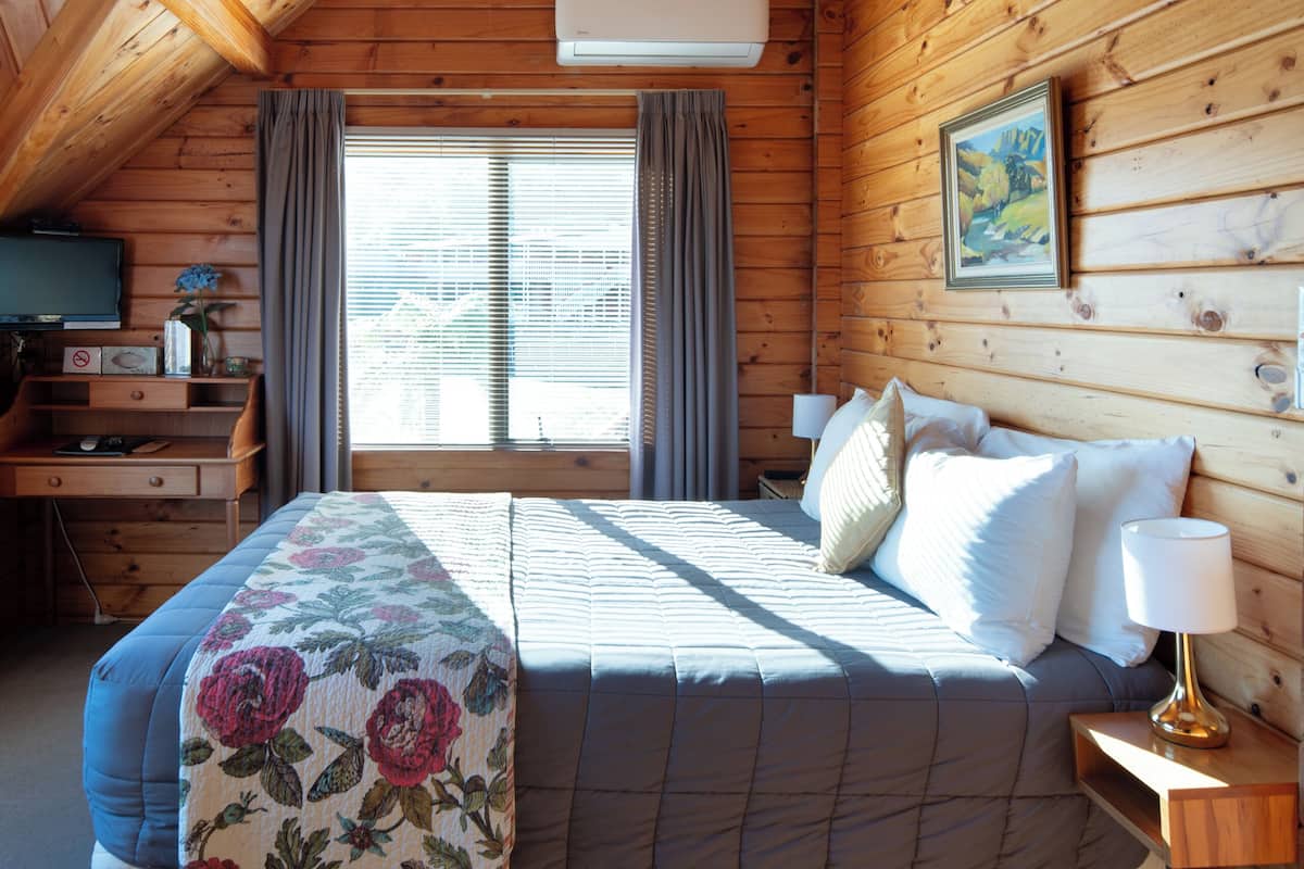 A cozy wooden cabin bedroom with a large bed, a nightstand, a lamp, a window with curtains, and a painting on the wall.