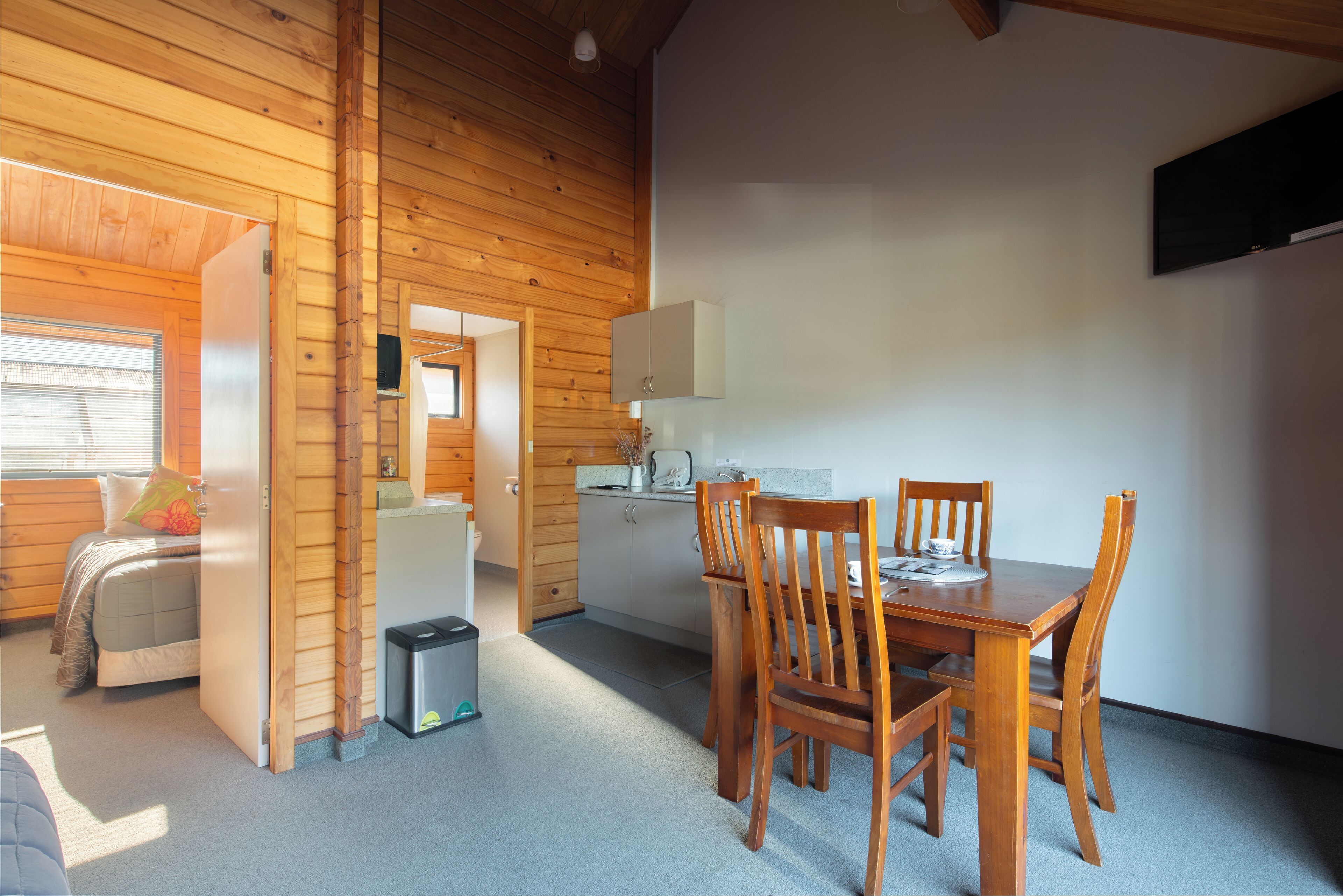 A compact room with a wooden ceiling, a bed, a small kitchenette, and a dining area with wooden chairs.