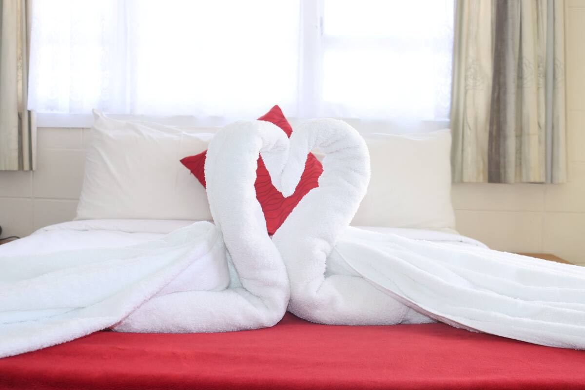 A bed with white and red bedding, a heart-shaped pillow, and a window with curtains.