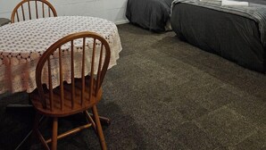 Una habitación con pared de ladrillo blanco, una mesa con mantel de encaje, dos camas individuales con ropa de cama oscura y una cortina roja.
