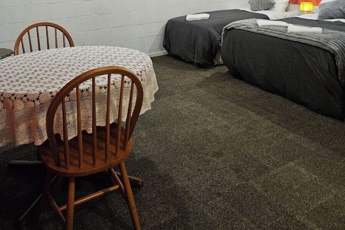 Quarto com parede de tijolos brancos, uma mesa com toalha rendada, duas camas de solteiro com roupa de cama escura e uma cortina vermelha.