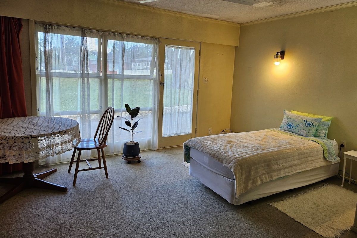 Quarto com cama, cadeira, mesa com toalha de renda, planta em vaso e janela com cortinas.