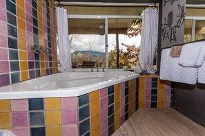 A bathroom with a large bathtub, colorful tiles, a bench, and a view of mountains through a glass door.