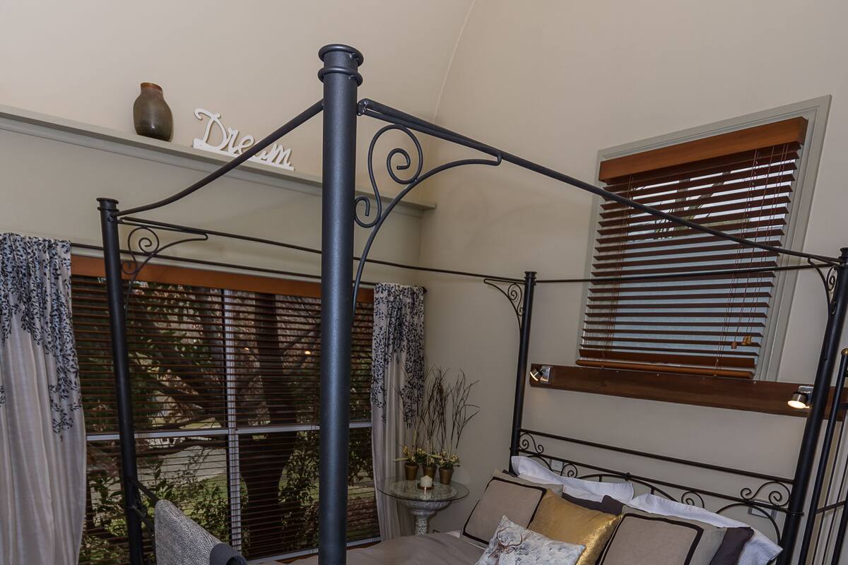 A four-poster bed with a grey bedspread and multiple pillows, a small bedside table with a vase, and a window with blinds.