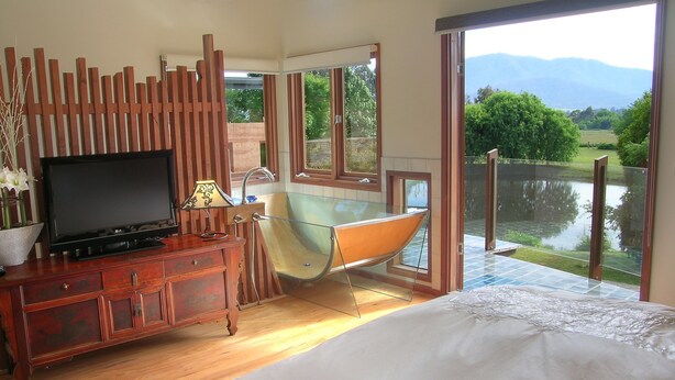 A bedroom with a large bed, a wooden dresser, a television, a glass-enclosed bathtub, and a view of a lake and mountains. - Dreamers (Tawonga South)